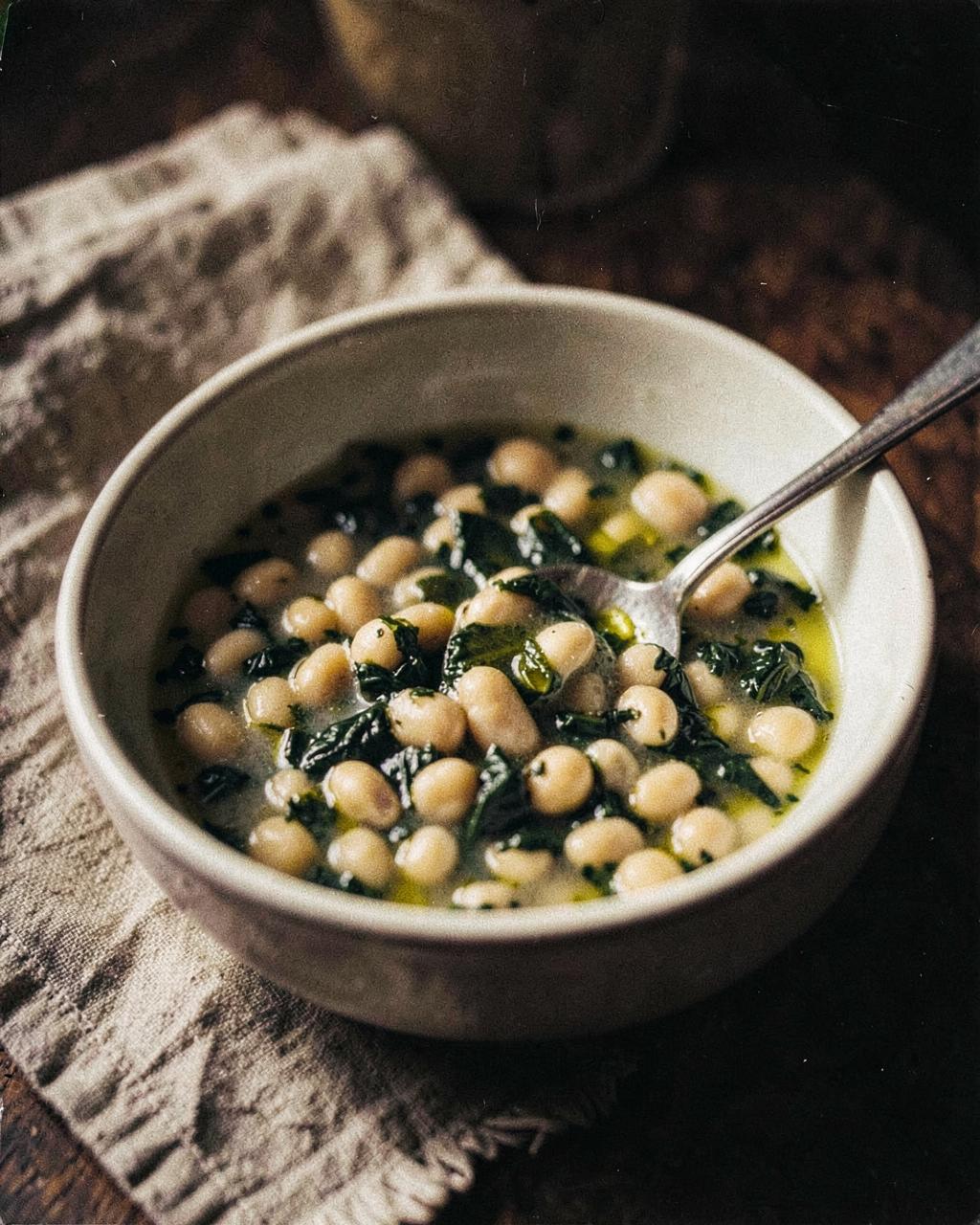 A bowl of brothy white beans with dark greens and olive oil, spoon resting inside
